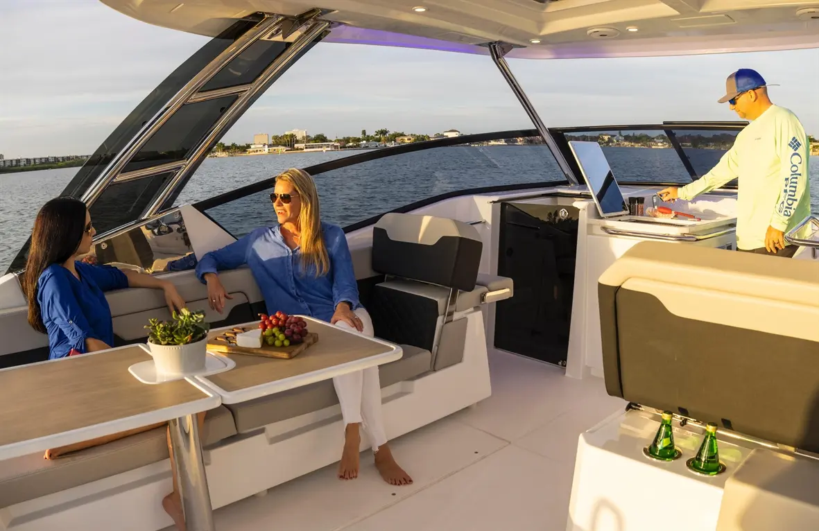 Two women in sunglasses relax on a boat, enjoying a platter of fruit and cheese while a man prepares food nearby.